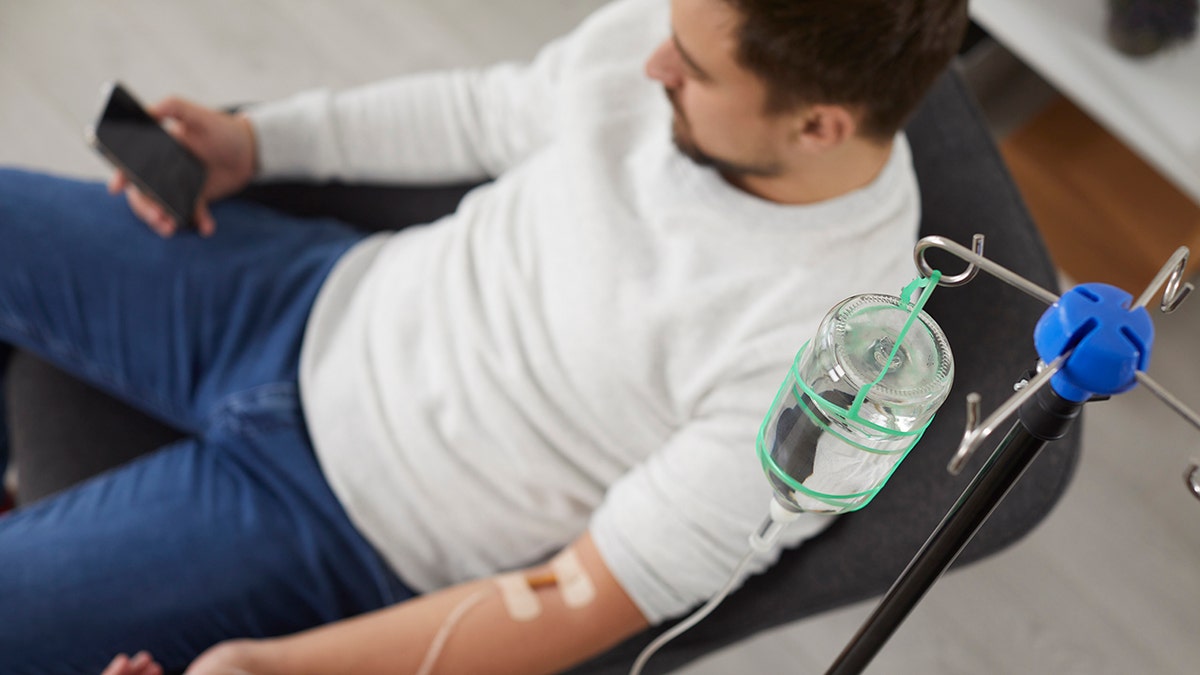 Higher view of a man sitting in the chair and using the smartphone while receiving infusion of Gotig IV and therapy of vitamins in his blood. Person who receives injection therapy. Health concept: used for cetamine crew